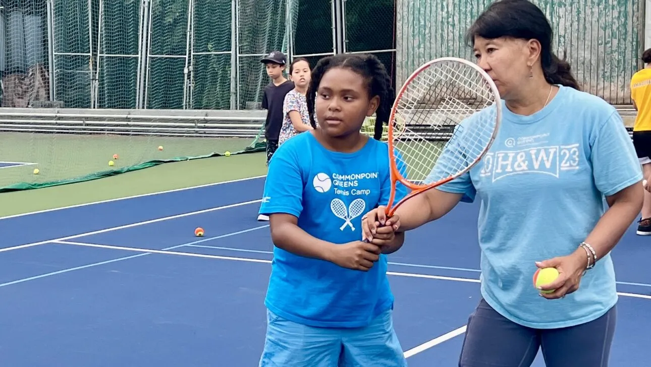 Ein Ausbilder leitet ein Kind mit einem Tennisschläger auf einem Außenplatz an, umgeben von Tennisbällen und anderen Teilnehmern im Hintergrund.