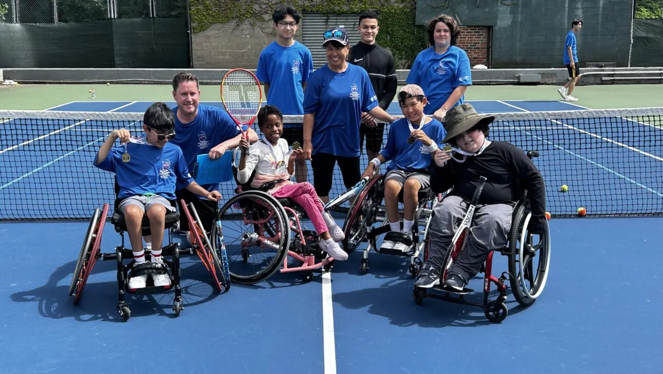 Eine Gruppe von Menschen, einige im Rollstuhl, lächelt und posiert auf einem Tennisplatz. Tennisschläger und Bälle sind sichtbar.