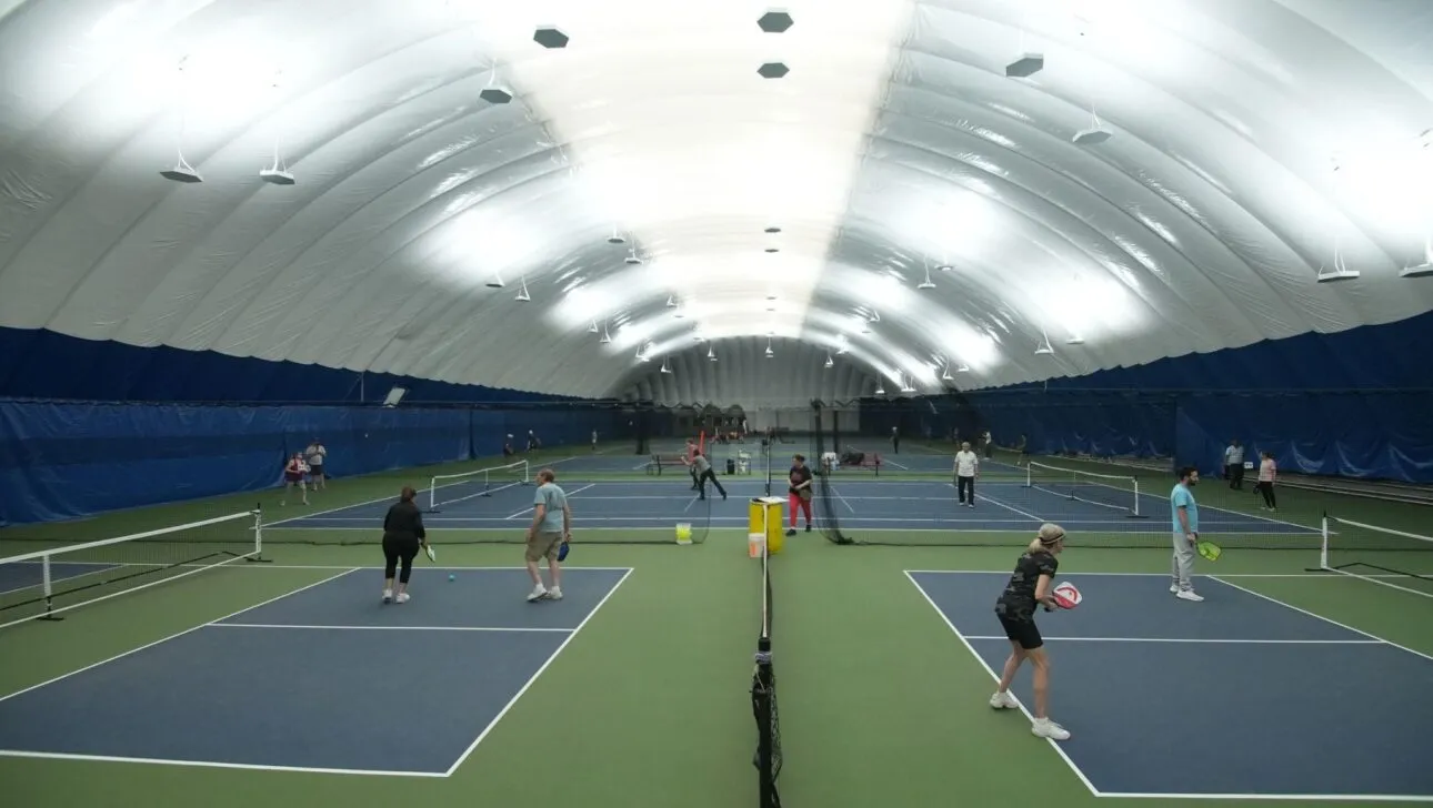 Innenanlage für Tennis mit mehreren Plätzen unter einer großen, weißen, gewölbten Decke.