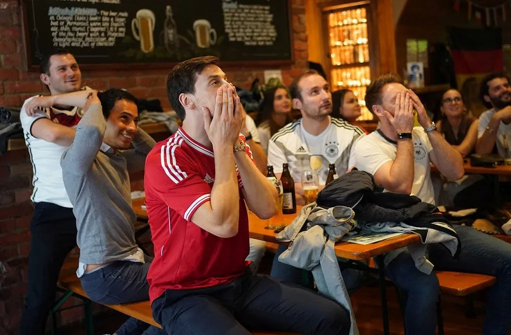 Junge Männer und vereinzelte Frauen teilweise in Trickots der deutschen Fußball-Nationalmannschaft sitzen in einem Pub und richten den Blick offenbar auf einen Bildschirm außerhalb des Bildes. Einige schlagen die Hände vors Gesicht oder über den Kopf.