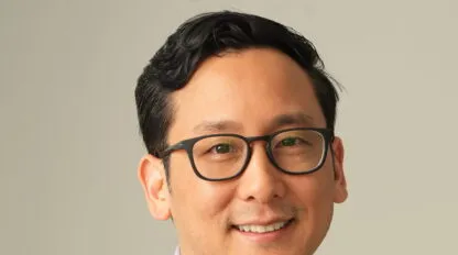 Mann mit kurzen schwarzen Haaren und Brille, trägt ein hellviolettes Hemd mit Knöpfen und lächelt vor einem schlichten Hintergrund.