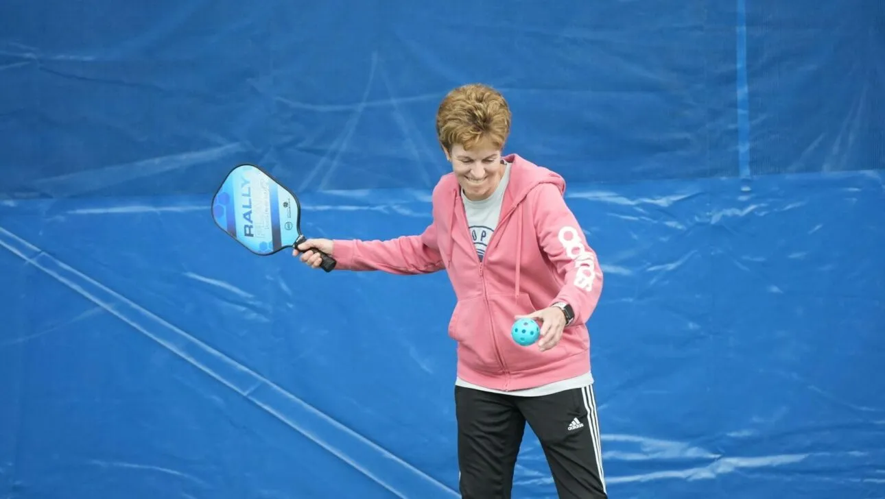Person in einem pinkfarbenen Hoodie bereitet sich darauf vor, einen Pickleball auf einem blauen Platz aufzuschlagen, und hält ein Paddel und einen Ball.