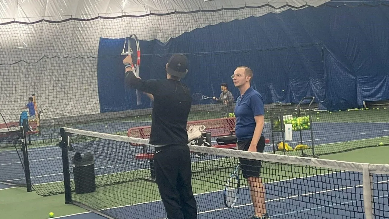Zwei Personen auf einem Hallen-Tennisplatz neben dem Netz; eine hält einen Schläger und zeigt ihn nach oben. Blaue und grüne Spielflächen sind sichtbar.