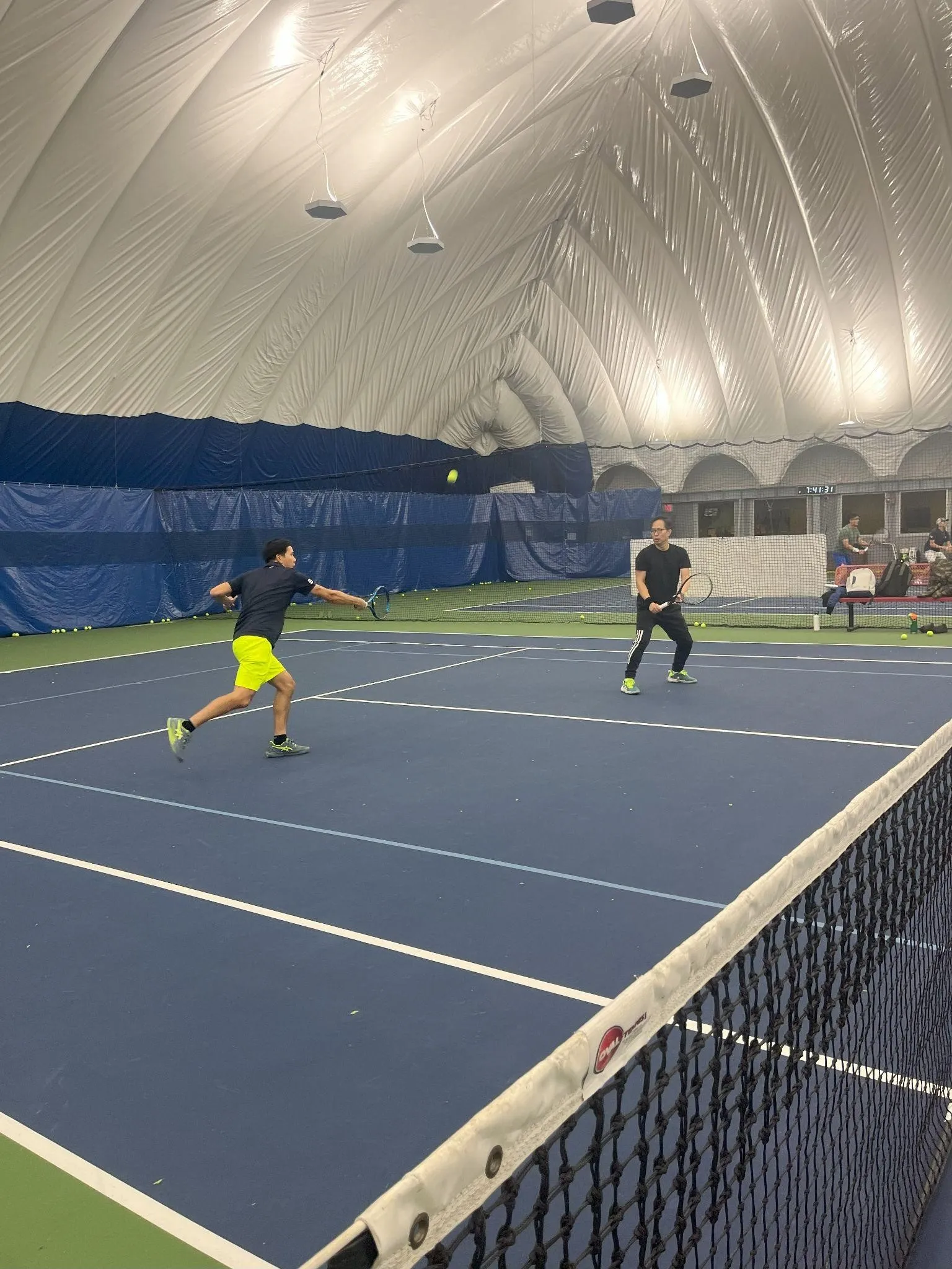Zwei Personen spielen Tennis drinnen auf einem blauen Platz. Eine Person bereitet sich darauf vor, den Ball mit einem Vorhandschwung zu treffen, während die andere in der Nähe des Netzes steht.