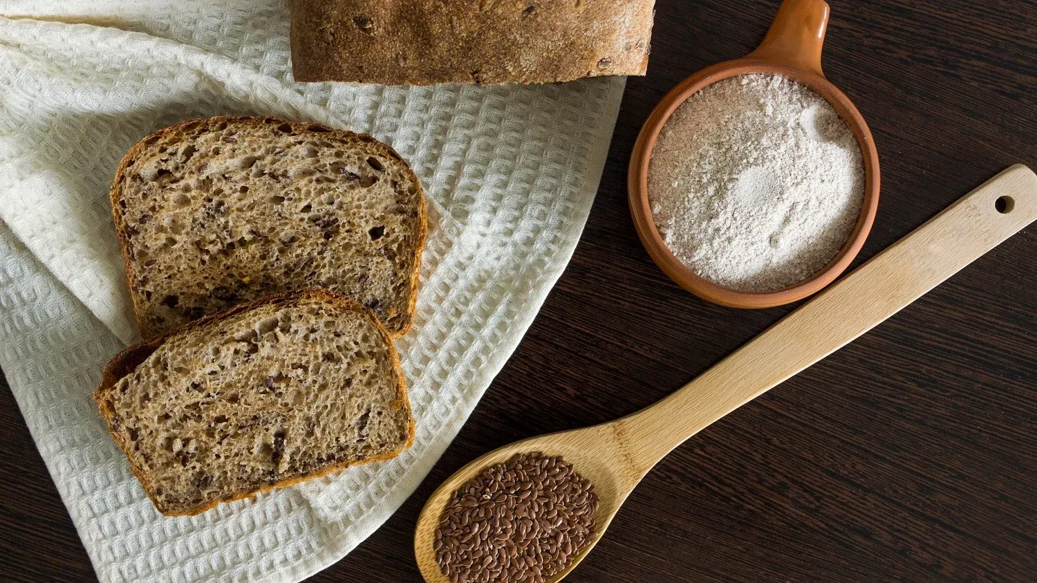Draufsicht: Auf einem dunklen Untergrund ist ein kleiner Teil eines Leinsamenbrotes zu sehen. Daneben liegen zwei Scheiben Brot, ein Holzlöffel mit leinsamen und eine Schüssel mit Mehl.