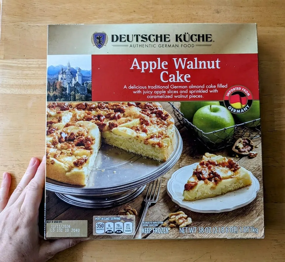 Eine Hand hält eine Schachtel Aldi Deutsche Küche Apfel-Walnuss-Kuchen auf einem Holztisch.