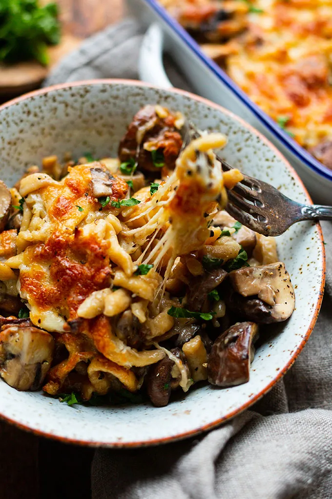 Eine Portion Spätzle-Pilz-Auflauf mit Käsefäden in einer Schüssel. Im Hintergrund sieht man die Auflaufform.