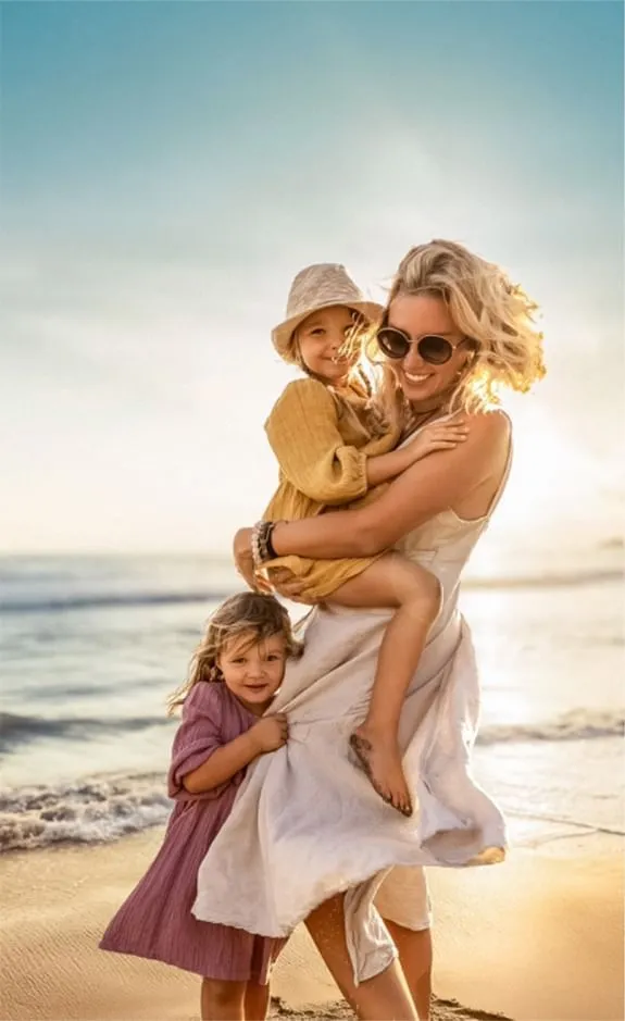 Mutter mit zwei Kindern am Strand, einer auf dem Arm, bei Sonnenuntergang.